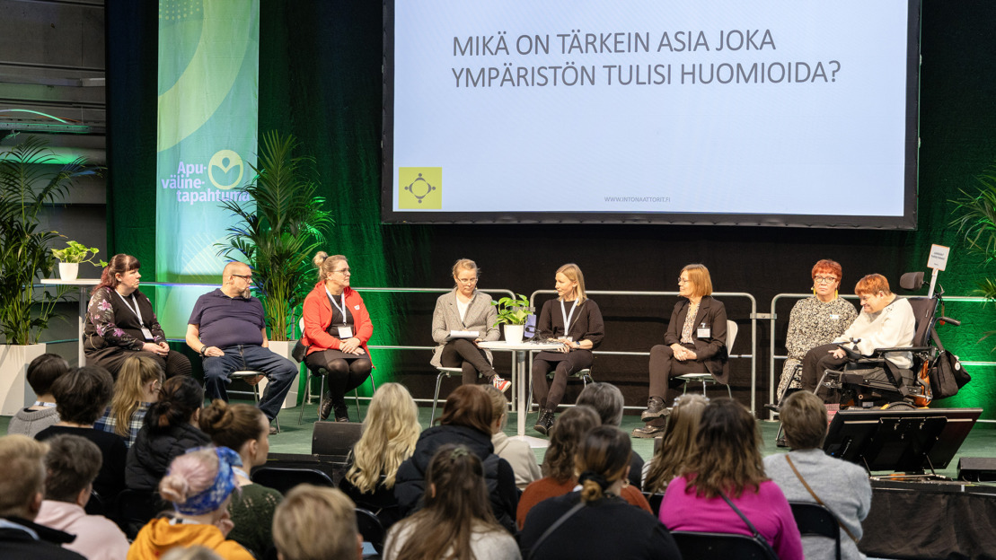 Kuva viime Apuvälinetapahtuman ohjelmasta. Lavalla menossa paneelikeskustelu, etualalla näkyy yleisöä, lavalla panelistien takana valkokankaalla teksti: Mikä on tärkein asia joka ympäristön tulisi huomioida?